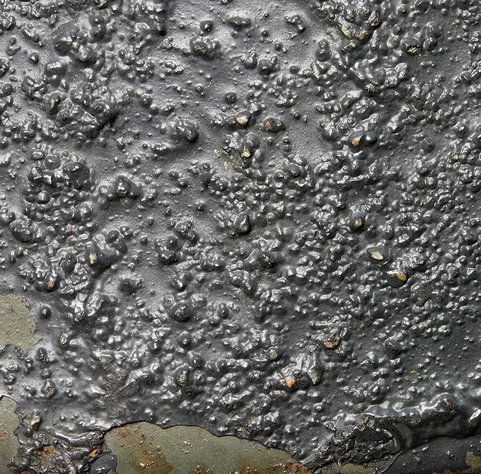 Nærbilde av malingsstrukturen på M35 hjelmen vist over. Legg merke til tykkelsen og inneholdet av småsteiner og sand. Man kan se Quist fabrikkmalingen hvor det tykke laget har falt av.